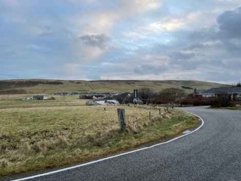 A winding road curves through a rural landscape with grassy fields, a small village in the distance, and partly cloudy skies overhead.