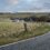 A winding road curves through a rural landscape with grassy fields, a small village in the distance, and partly cloudy skies overhead.