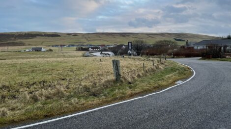 A winding road curves through a rural landscape with grassy fields, a small village in the distance, and partly cloudy skies overhead.