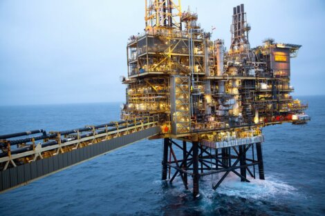 An offshore oil rig stands tall in the ocean, connected by a bridge. The structure is illuminated, highlighting intricate machinery against a backdrop of cloudy skies and calm waters.
