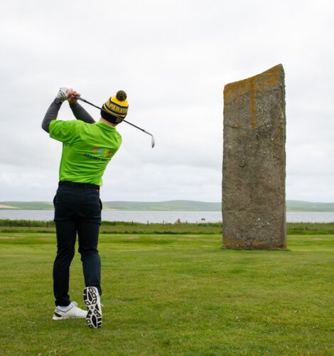 Golfer in a green shirt and knit hat swings a club near a large vertical stone on a grassy field, with water and hills in the background.