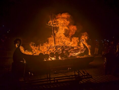 A Viking-style ship engulfed in flames during a nighttime event, with onlookers silhouetted against the fire.
