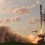 A rocket launches vertically from a launchpad, producing flames and thick smoke, with a partly cloudy sky in the background.