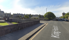 Street view of King Harald Street with stone buildings, a paved sidewalk, and parked cars on the right. Green lawns and trees line the road under a clear sky.