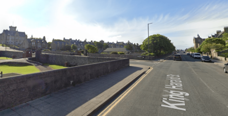 Street view of King Harald Street with stone buildings, a paved sidewalk, and parked cars on the right. Green lawns and trees line the road under a clear sky.