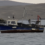 A small blue and white fishing boat is anchored near a rocky shoreline, with hilly terrain in the background.