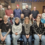 A group of fifteen people, consisting of men and women, pose indoors in two rows; some are seated in front while others stand behind them, all facing the camera.