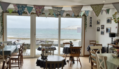 Cozy cafe with wooden chairs and tables, each covered with dotted tablecloths. Bunting hangs from the ceiling. Large windows offer a view of the ocean and snowy landscape outside.