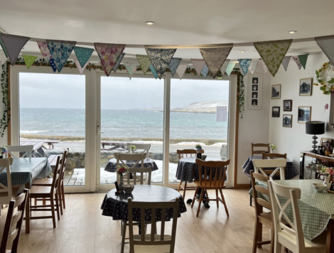 Cozy cafe with wooden chairs and tables, each covered with dotted tablecloths. Bunting hangs from the ceiling. Large windows offer a view of the ocean and snowy landscape outside.