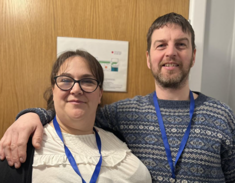 Two people wearing lanyards stand side by side in front of a wooden door. One person has an arm around the other. They are smiling and wearing casual clothing.