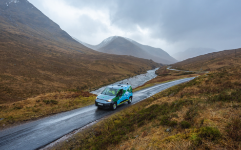 A van drives along a narrow road in a mountainous landscape, with a river running parallel in overcast weather.
