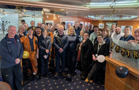 A large group of people gathered indoors celebrating a retirement, with balloons and a banner in the background.