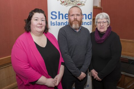 Three people standing in front of a Shetland Islands Council banner inside a room with wood-paneled walls.