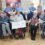A group of elderly people, some in wheelchairs, sit and stand with caregivers in a care home, holding a large charity check and smiling for the camera.