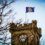 Flag flying atop a stone clock tower, with tree branches in the foreground and a clear sky.