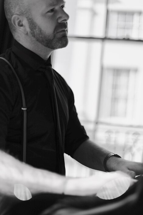 Black and white photo of a man with a beard wearing a dark shirt and tie, standing near a window.