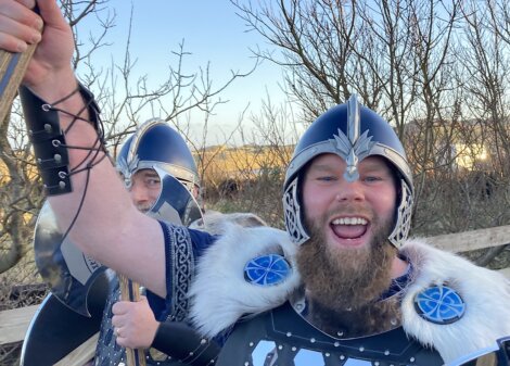 Two people in Viking-style armor, one in front smiling broadly, holding a pole, with trees and blue sky in the background.