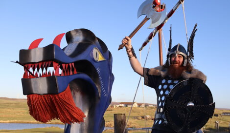 Person in Viking costume with an axe stands next to a dragon ship's head outdoors.