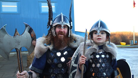 Two people dressed in Viking costumes, one adult and one child, wearing helmets and fur-trimmed cloaks, holding an axe and a spear, standing outside a blue building.