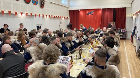A group of people in fur-trimmed outfits sit at long tables, eating and reading booklets, in a decorated hall.