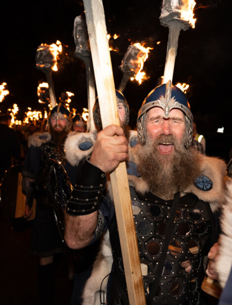 A bearded warrior in Viking armor holds a torch, yelling energetically. Other torch-bearing figures are visible in the background.