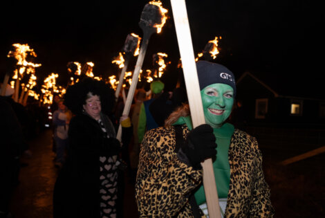 People with painted faces and costumes hold flaming torches during a nighttime parade.
