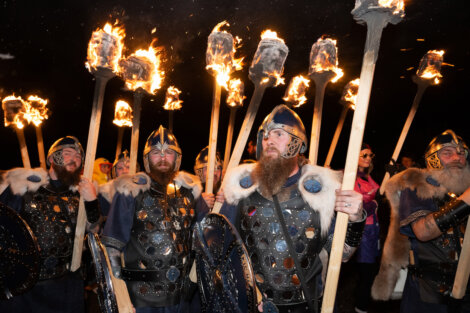 Group of people in Viking costumes holding flaming torches at night, dressed in helmets, fur, and armor.