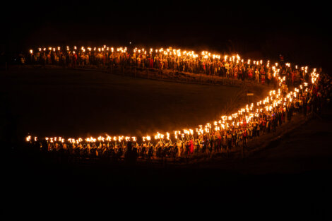 A large group of people holding torches form a circular shape in the dark, creating a bright ring of light against the night.