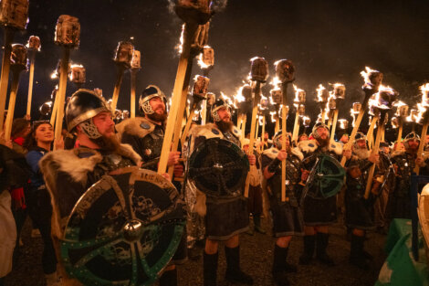 Group of people in medieval-style costumes holding large flaming torches and shields during a night event.