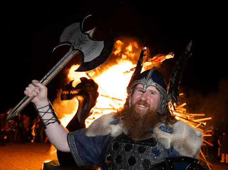 Person in Viking attire holds an axe and smiles in front of a burning structure at night.