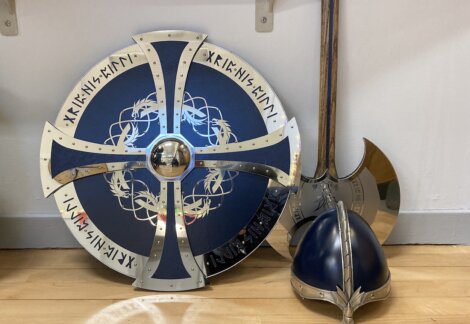 A round shield with runes and a dragon design, an axe with a metallic blade, and a blue helmet with a metallic crest resting on a wooden floor.
