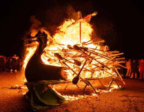 A wooden Viking ship replica is engulfed in flames during a nighttime event, with onlookers visible in the background.