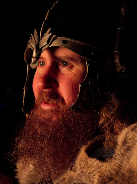 A man with a long beard is wearing a metal helmet with intricate designs, looking to the side. The lighting casts shadows on his face, enhancing the helmet's details.