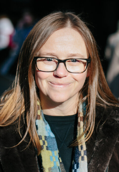 A woman with long brown hair and glasses, wearing a patterned scarf, smiles at the camera.