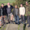 Five men standing on a stone path outdoors, one wearing camo pants, another in a camo jacket, with trees and a field in the background.