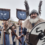 A man in Viking-style costume, holding an axe and shield with a dragon emblem, stands in front of a group of similarly dressed people carrying banners.