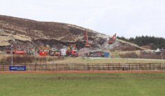 Construction site with several excavators, trucks, and equipment working on a hillside. A grassy field and a small building are visible in the foreground.