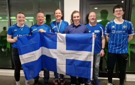 Six people standing indoors, smiling and holding a large blue and white flag with a cross design. They are wearing various blue tops.