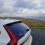 A white Volvo car with rear lights on is parked on a coastal road, overlooking grassy fields and the sea under a cloudy sky.