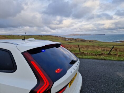 A white Volvo car with rear lights on is parked on a coastal road, overlooking grassy fields and the sea under a cloudy sky.