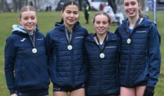 Four athletes wearing medals stand together outdoors, dressed in navy sports jackets and shorts, smiling for a group photo.