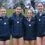 Four athletes wearing medals stand together outdoors, dressed in navy sports jackets and shorts, smiling for a group photo.