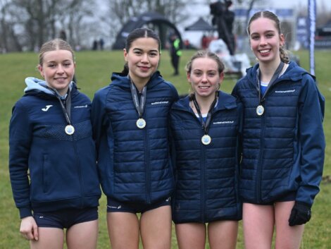 Four athletes wearing medals stand together outdoors, dressed in navy sports jackets and shorts, smiling for a group photo.