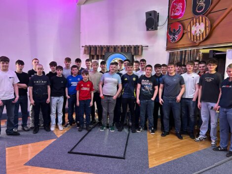 A group of men and boys stand together in a room, posing for a photo. Some are wearing casual t-shirts, others have shirts with logos. There's a clock and decorations on the wall behind them.