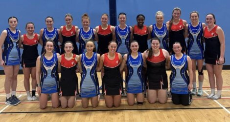 A group of women in team uniforms pose for a photo in a sports hall, with a mix of red and blue designs on their outfits.