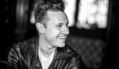 Black and white image of a smiling person with short hair, wearing a leather jacket and a light-colored shirt, in front of a blurred background.