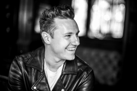 Black and white image of a smiling person with short hair, wearing a leather jacket and a light-colored shirt, in front of a blurred background.
