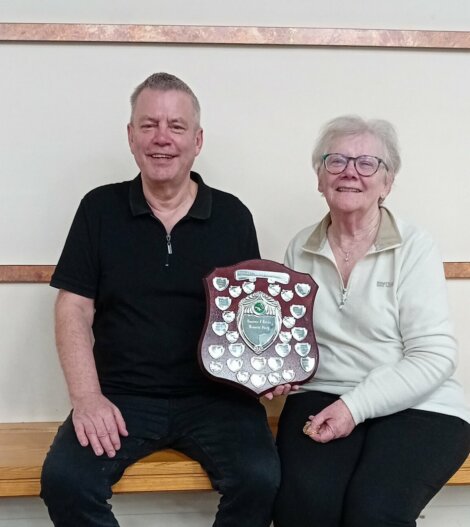 Two people sitting on a bench holding a shield-shaped award with multiple small plaques.