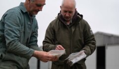 Two men examining samples in clear plastic bags outdoors, standing near industrial buildings.
