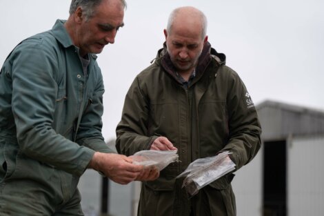Two men examining samples in clear plastic bags outdoors, standing near industrial buildings.